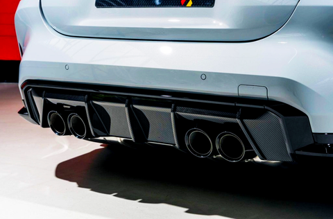 A white car features quad exhaust pipes integrated into a black carbon fiber diffuser, displayed in a well-lit indoor setting with a red wall in the background.