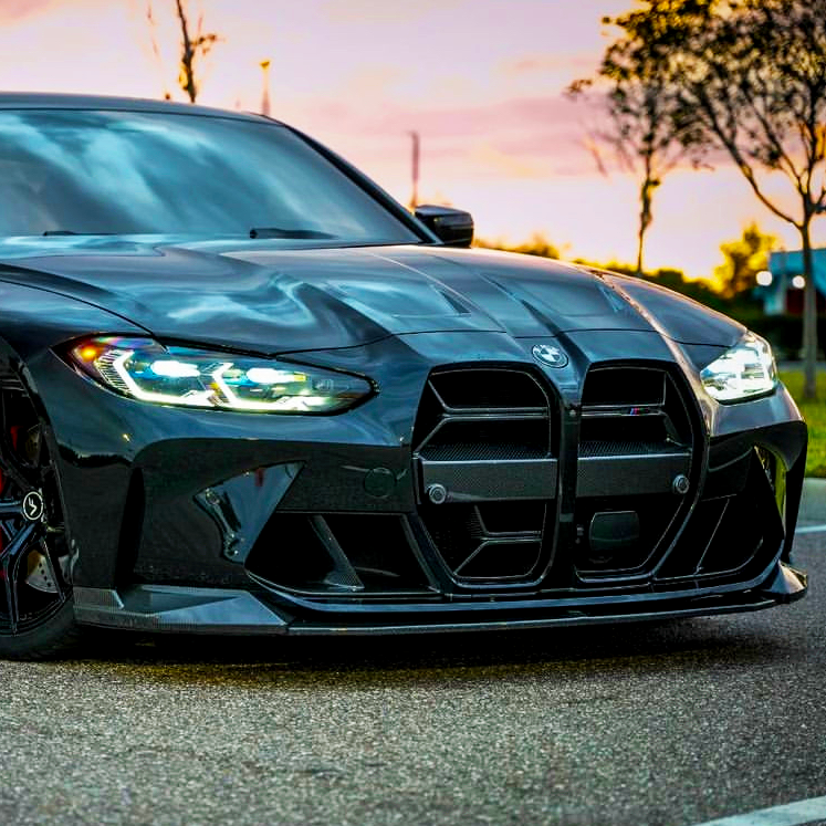 A sleek, glossy black sports car is parked on a paved road at sunset. Its prominent grille and illuminated headlights are emphasized against a backdrop of trees and a colorful sky.