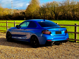 A blue sports coupe is parked on a gravel path, surrounded by a wooden fence and green field, with trees in the background under a partly cloudy sky.