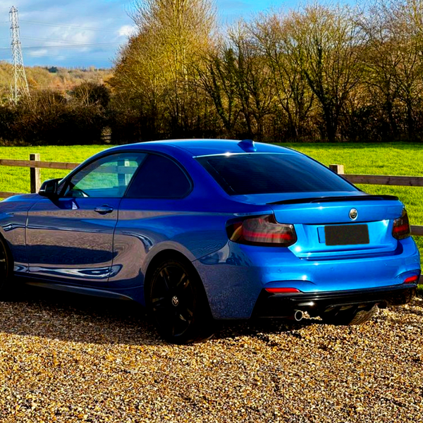 A blue sports coupe is parked on a gravel path, surrounded by a wooden fence and green field, with trees in the background under a partly cloudy sky.