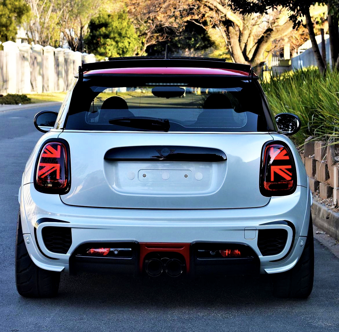 A white car is parked on a residential street. The rear features distinctive Union Jack tail lights, with a red roof spoiler. Trees and greenery line the background.