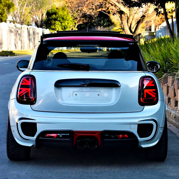 A white car is parked on a residential street. The rear features distinctive Union Jack tail lights, with a red roof spoiler. Trees and greenery line the background.