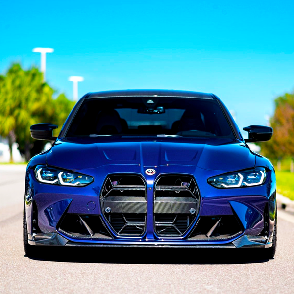 A sleek blue car is stationary on a sunny road, with its distinctive large grille and angular headlights prominently displayed, surrounded by blurred greenery and a clear blue sky.