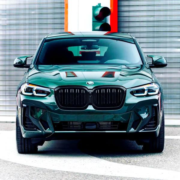 A green BMW car is parked on a circular pavement, positioned in front of a brushed metal wall and a traffic light.