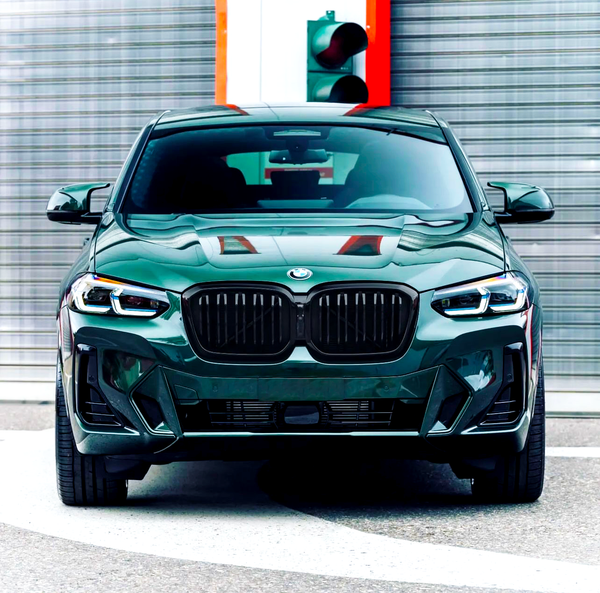 A green BMW car is parked on a circular pavement, positioned in front of a brushed metal wall and a traffic light.