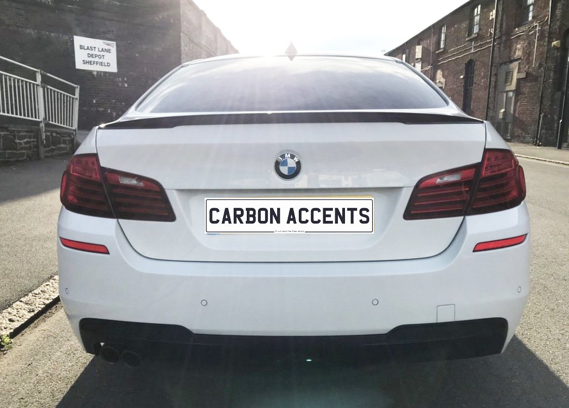 A white BMW sedan is parked on a street, featuring a license plate reading "CARBON ACCENTS." In the background, there's a building with a sign saying "BLAST LANE DEPOT SHEFFIELD."