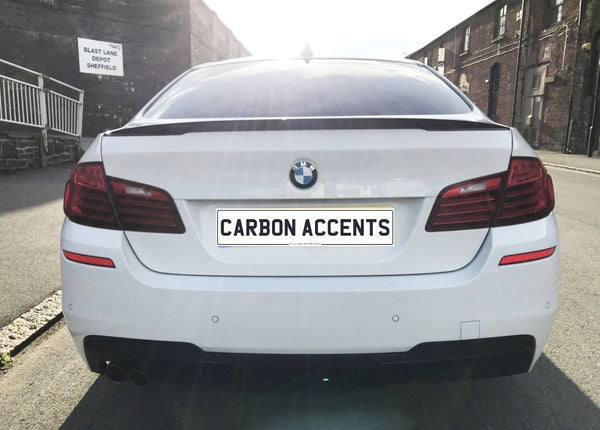 A white BMW sedan is parked on a street, featuring a license plate reading "CARBON ACCENTS." In the background, there's a building with a sign saying "BLAST LANE DEPOT SHEFFIELD."