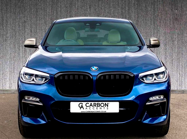 A blue BMW car is parked on pavement, with a textured wall backdrop. The license plate reads “CARBON ACCENTS www.CARBONACCENTS.co.uk.”