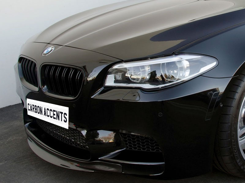 A black car with a BMW badge is parked on a dark surface, featuring prominent headlights and a "CARBON ACCENTS" license plate. The background is a plain, light-colored wall.