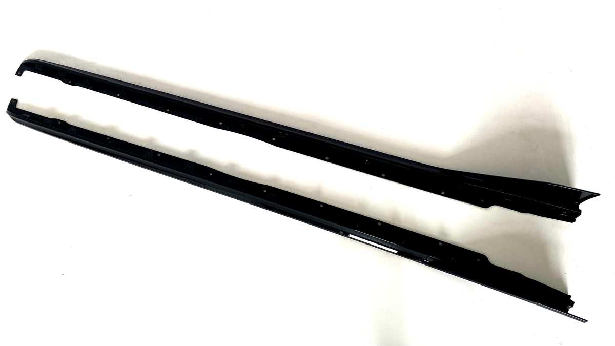 Two elongated black metal car side skirts lie parallel on a plain white surface, featuring multiple mounting holes along their length and slight contours for aerodynamic enhancement.