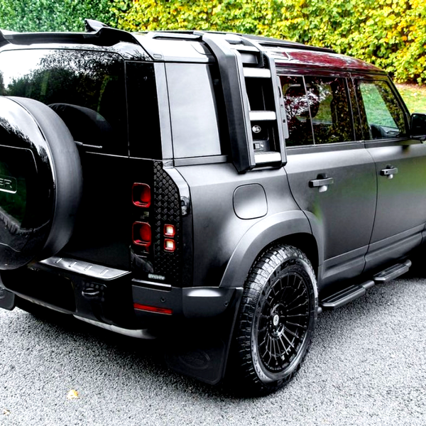 SUV with a spare tire labeled "DEFENDER" is parked on a paved road, surrounded by lush green grass and foliage in the background.