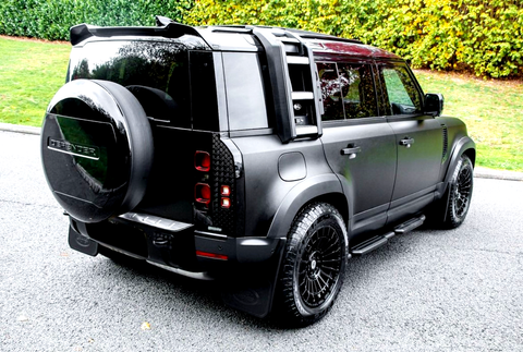 SUV with a spare tire labeled "DEFENDER" is parked on a paved road, surrounded by lush green grass and foliage in the background.