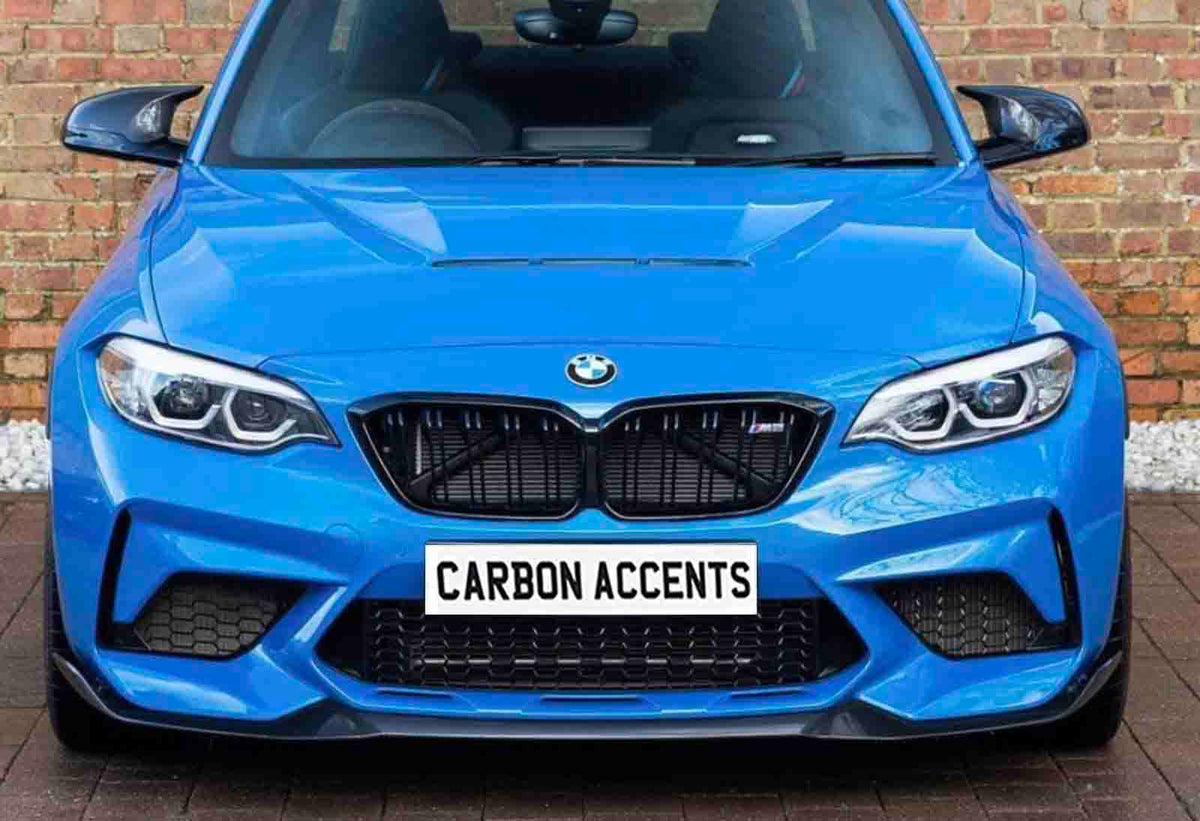 A bright blue BMW car is parked facing forward, displaying a bold front grille and headlights. The license plate reads "CARBON ACCENTS." It is set against a brick wall backdrop.