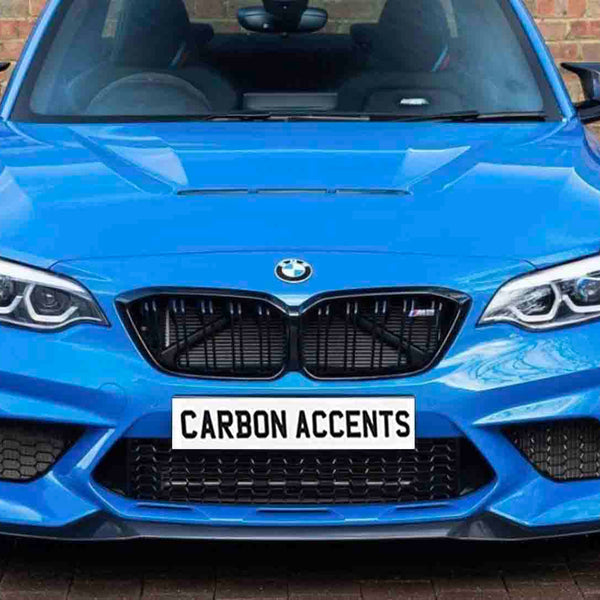 A bright blue BMW car is parked facing forward, displaying a bold front grille and headlights. The license plate reads "CARBON ACCENTS." It is set against a brick wall backdrop.