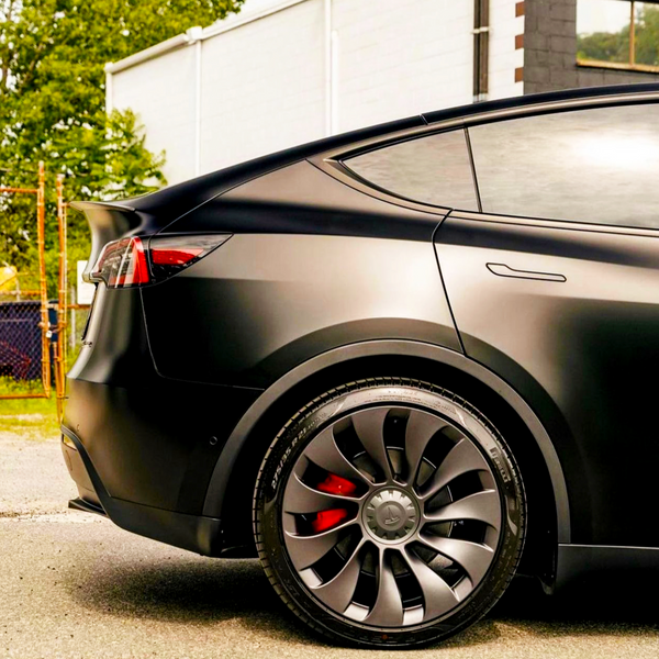 A sleek, black car features aerodynamic bodywork and modern wheels. It is parked outdoors on a paved surface next to a white building and a tree.