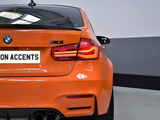 An orange BMW M3 is parked indoors, highlighting its rear view with illuminated taillights. The license plate reads 