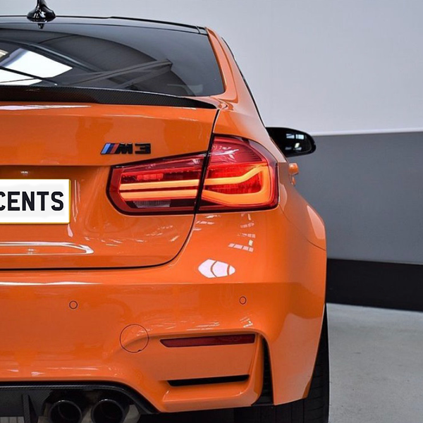 An orange BMW M3 is parked indoors, highlighting its rear view with illuminated taillights. The license plate reads "CARBON ACCENTS," and the environment features a grey wall and concrete floor.