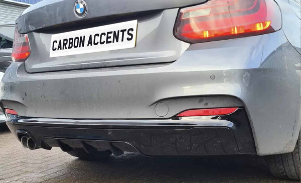 A silver car displays a "CARBON ACCENTS" license plate, featuring dual exhaust pipes and red taillights. It is parked on a textured surface, with a building in the background.