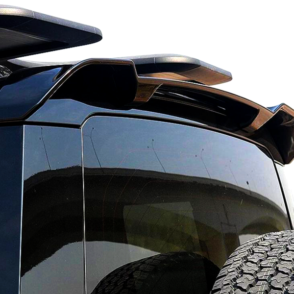 A black SUV with a rooftop spoiler and a rear tire mounted on the back, parked outdoors on a sunny day, with a partial view of a curved overpass in the background.