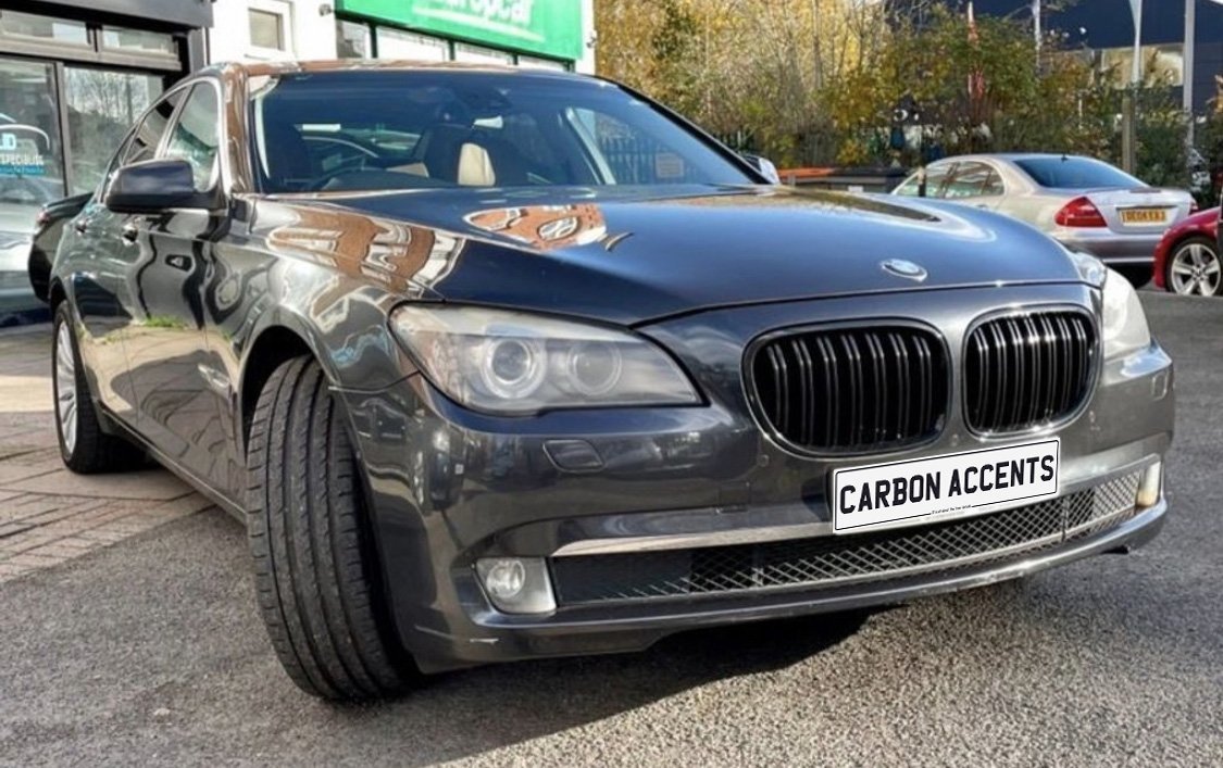 Car parked outside a building with "CARBON ACCENTS" on the license plate, surrounded by other vehicles and trees. The car has a glossy, dark finish and prominent front grille.