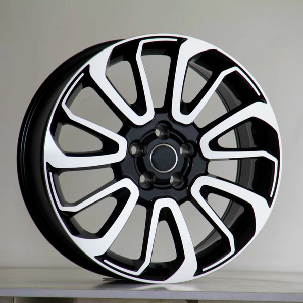 A black and white alloy wheel with ten curved spokes sits against a plain, light-gray background, resting on a flat surface.