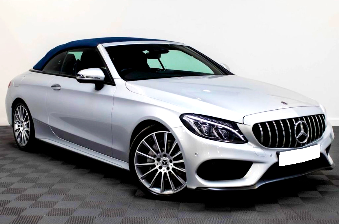 Silver convertible car, parked in an indoor showroom. The vehicle features a blue soft-top roof, sleek body lines, and distinctive front grille with a logo. The floor is checkered tile.