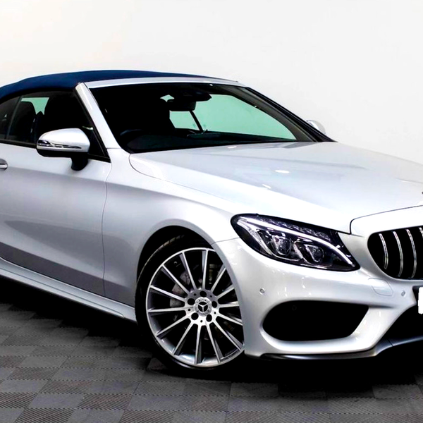 Silver convertible car, parked in an indoor showroom. The vehicle features a blue soft-top roof, sleek body lines, and distinctive front grille with a logo. The floor is checkered tile.