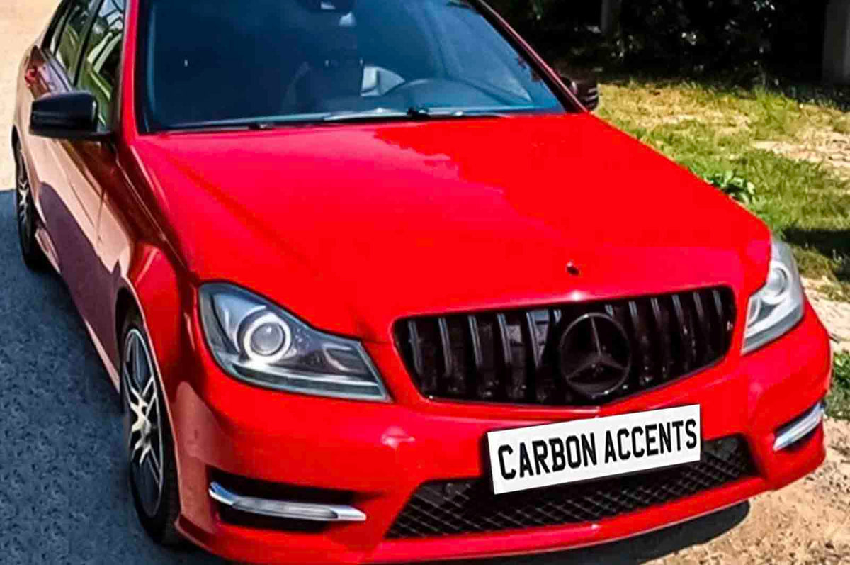 Red car with a black grille and prominent headlights, parked on a road beside a grassy area. The license plate reads "CARBON ACCENTS."