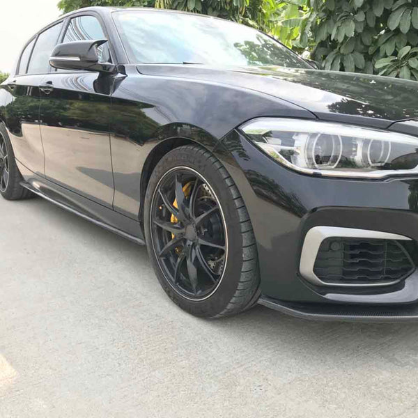 A sleek black car with sporty wheels and yellow calipers is parked on a concrete surface, surrounded by lush green foliage and trees in the background.