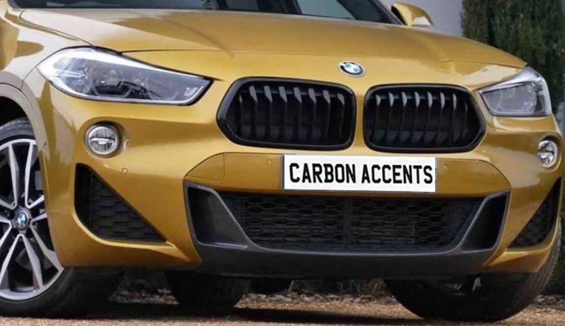 A gold-colored car front with distinctively styled headlights and grille. The license plate reads "CARBON ACCENTS." The setting appears to be outdoors, possibly near a building.
