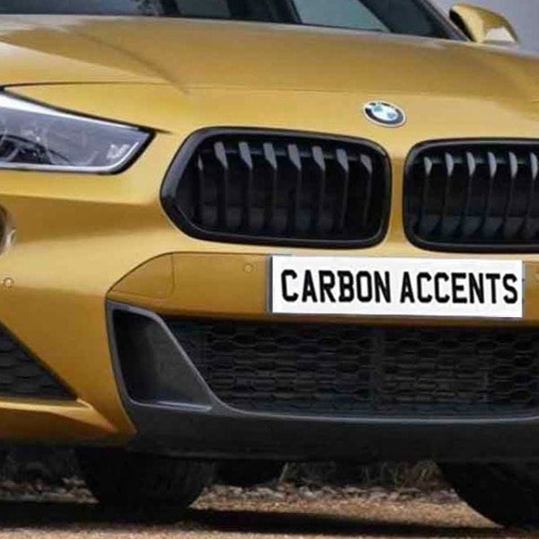 A gold-colored car front with distinctively styled headlights and grille. The license plate reads "CARBON ACCENTS." The setting appears to be outdoors, possibly near a building.