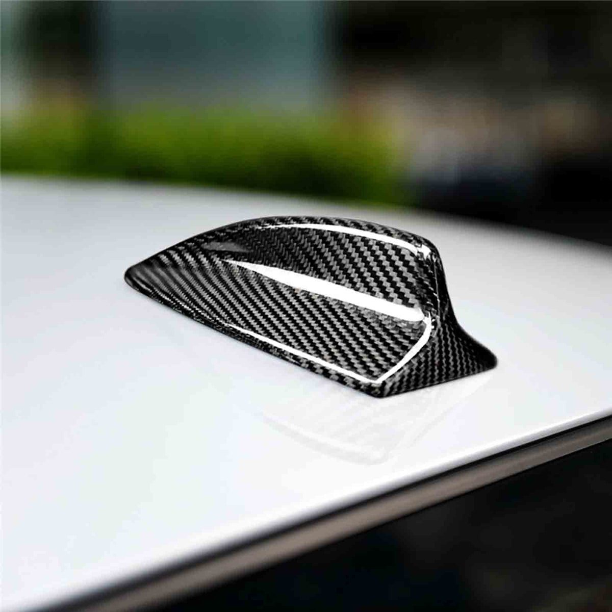 Carbon fiber shark fin antenna is mounted on a smooth, glossy white car roof, with a blurred green and gray background, suggesting an outdoor setting. No text present.