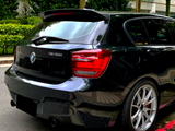 A black BMW M135i is parked on a street beside a hedge. The taillights glow, and the polished silver wheels stand out against the green foliage behind.