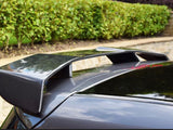 A carbon fiber car spoiler is mounted on the rear of a dark vehicle, surrounded by a neatly trimmed hedge and stone wall in a garden setting.