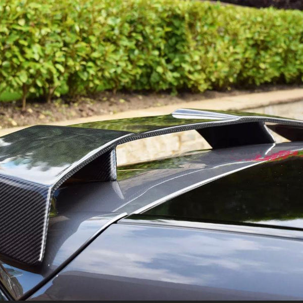A carbon fiber car spoiler is mounted on the rear of a dark vehicle, surrounded by a neatly trimmed hedge and stone wall in a garden setting.