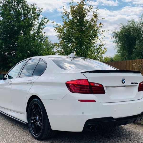 A white BMW M Sport sedan is parked on a gravel road beside a wooden fence, surrounded by trees under a partly cloudy sky.