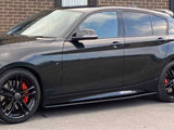 A shiny black car, parked on a street, displays red brake calipers and black alloy wheels. It is beside a brick building with large windows.