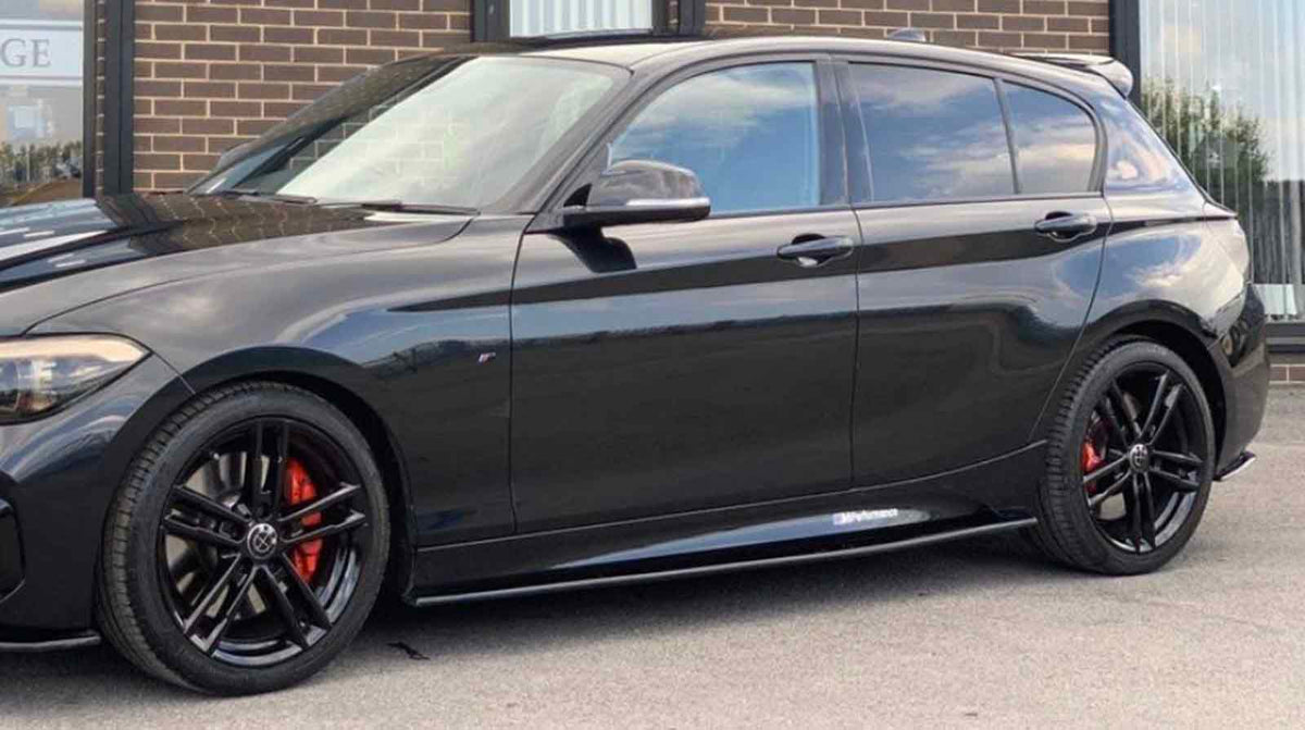 A shiny black car, parked on a street, displays red brake calipers and black alloy wheels. It is beside a brick building with large windows.