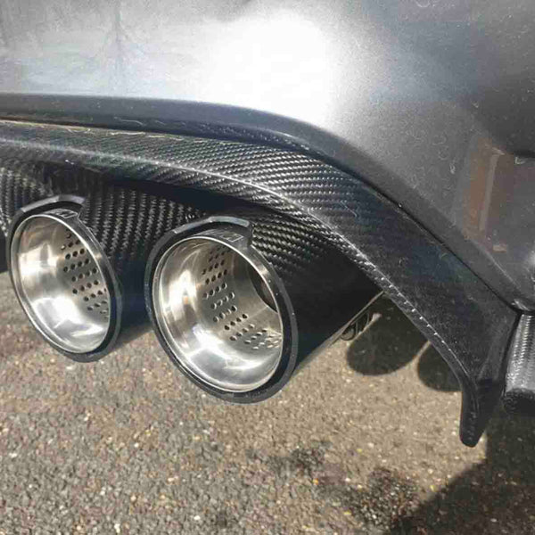 Car exhaust pipes made of shiny metal protrude from a carbon fiber rear diffuser, positioned above a gravel surface, with sunlight reflecting off the vehicle's body.
