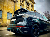 A sleek black car parked on a street in front of a neoclassical building with tall columns and a pediment. Sky is overcast, adding to the dramatic atmosphere.