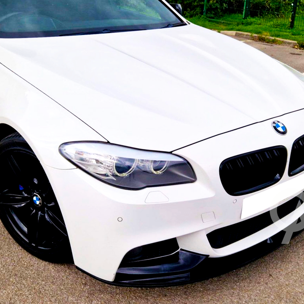 White BMW car, parked on asphalt, featuring black rims and a prominent front grille. It's situated in a parking lot bordered by green grass and a wire fence.