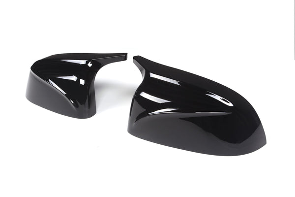 Two glossy black side mirror covers lying on a white surface. They are sleek, aerodynamic in design, and positioned at an angle to show their contours.