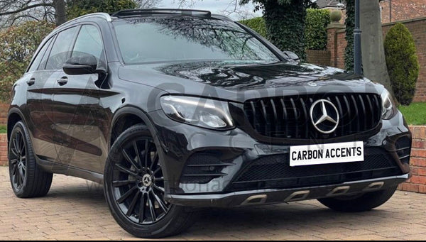 A black SUV is parked on a brick driveway. The vehicle features a prominent grille and black alloy wheels. Trees and a brick post are visible in the background. License plate reads "CARBON ACCENTS".