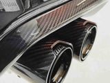 Exhaust pipes with a carbon fiber finish protruding from a vehicle's rear bumper, set in a brightly lit garage.