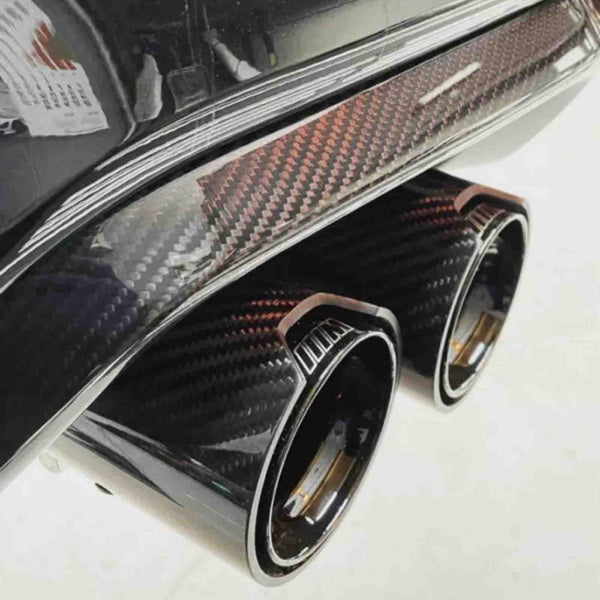 Exhaust pipes with a carbon fiber finish protruding from a vehicle's rear bumper, set in a brightly lit garage.