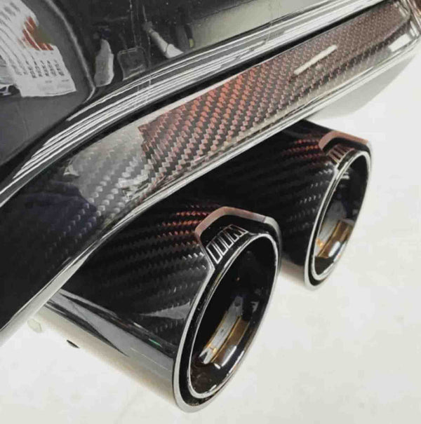 Exhaust pipes with a carbon fiber finish protruding from a vehicle's rear bumper, set in a brightly lit garage.