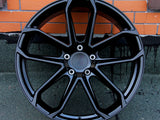 A black, multi-spoke car wheel rim is leaning against an orange brick wall, resting on a dark, textured surface.