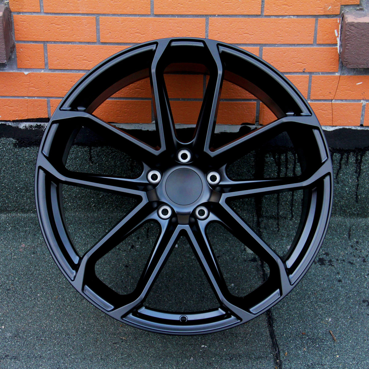 A black, multi-spoke car wheel rim is leaning against an orange brick wall, resting on a dark, textured surface.