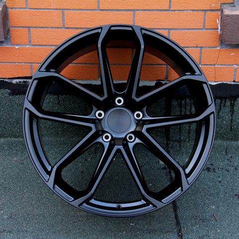 A black, multi-spoke car wheel rim is leaning against an orange brick wall, resting on a dark, textured surface.