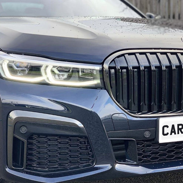 Car features illuminated headlights and a prominent grille. The bright wheels enhance its sleek design. Text reads "CARBON ACCE…" on the plate. The car is parked on asphalt beside a wooden fence.